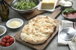 A pinsa crust on a cutting board surrounded by ingredients.